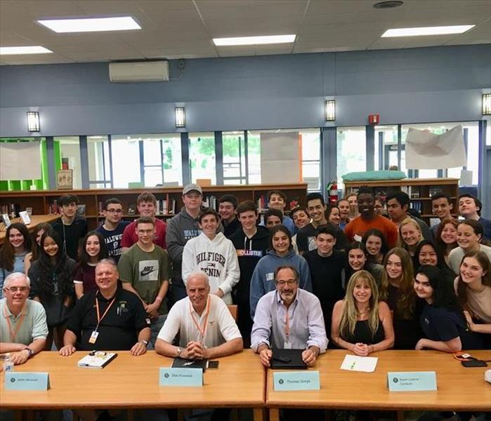 Group picture of high school students, and adults sitting at a table in front of them. 